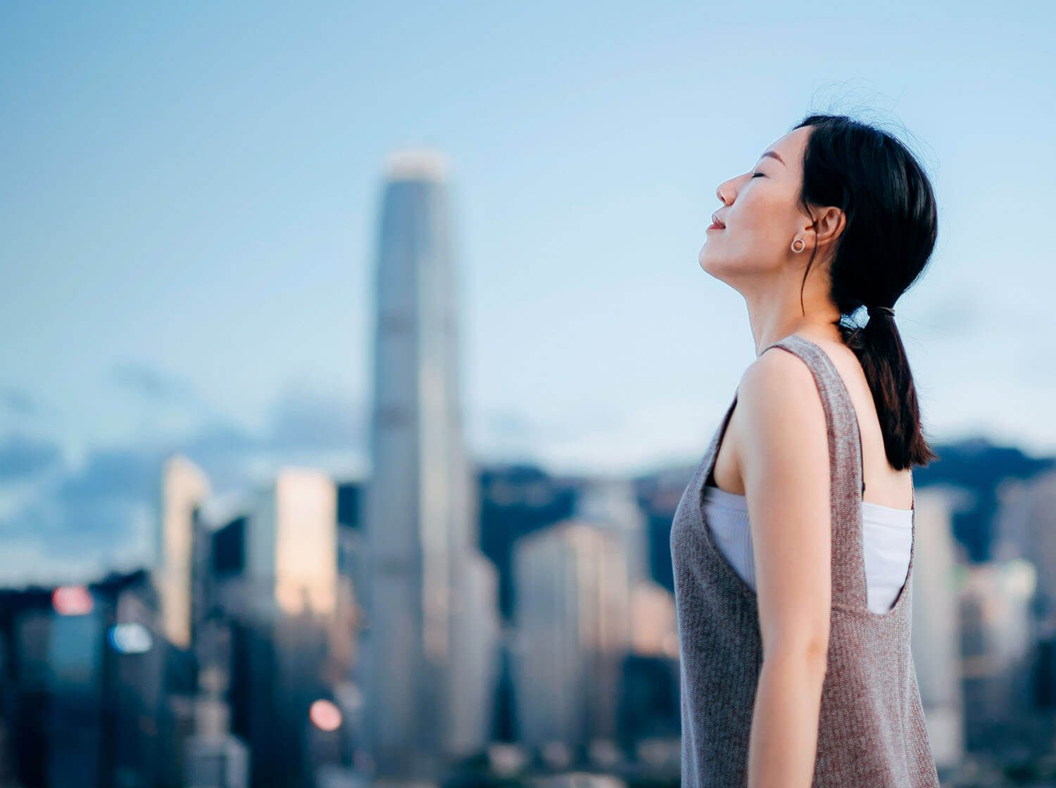 Against the backdrop of a city panorama, a woman raises her head with her eyes closed.